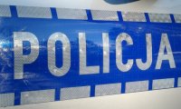 Fotografia kolorowa przedstawiająca srebrny napis policja na niebieskim tle. Umiejscowiony na bocznych drzwiach radiowozu