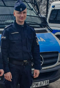 Fotografia kolorowa przedstawiająca policjanta stojącego przed radiowozem