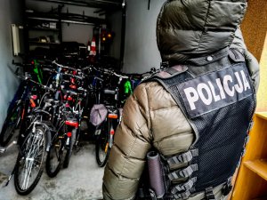 Policjant w ubraniach cywilnych, z założoną czarną kamizelką z napisem „POLICJA”. 
Policjant stoi przed garażem, w którym ustawione są skradzione, odzyskane rowery.