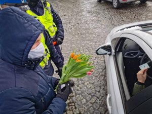 dwóch policjantów białostockiej drogówki oraz pracownik WORD, którzy stoją przy kontrolowanym srebrnym pojeździe. Pracownik trzyma bukiet tulipanów i jednego z nich wręcza kobiecie