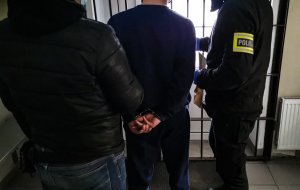 policjanci w cywilnych ubraniach trzymają zatrzymanego mężczyznę. Jeden z policjantów ma założoną żółtą, odblaskową opaskę z napisem &quot;POLICJA&quot;